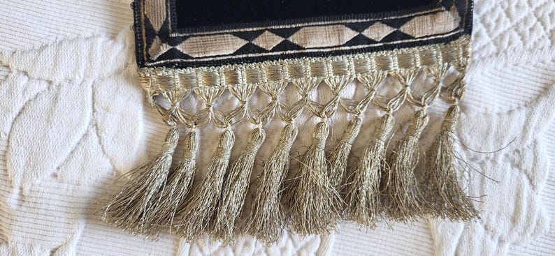 May include: Close-up of a decorative textile with a black velvet panel and a geometric pattern in gold and beige. The bottom edge features a fringe of gold tassels, adding a touch of elegance. The textile is likely used for home decor.