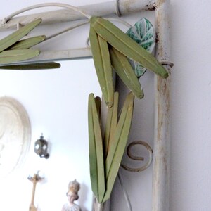 May include: A white metal mirror with a decorative design of green leaves. The mirror has a distressed white finish and is framed with a metal frame.