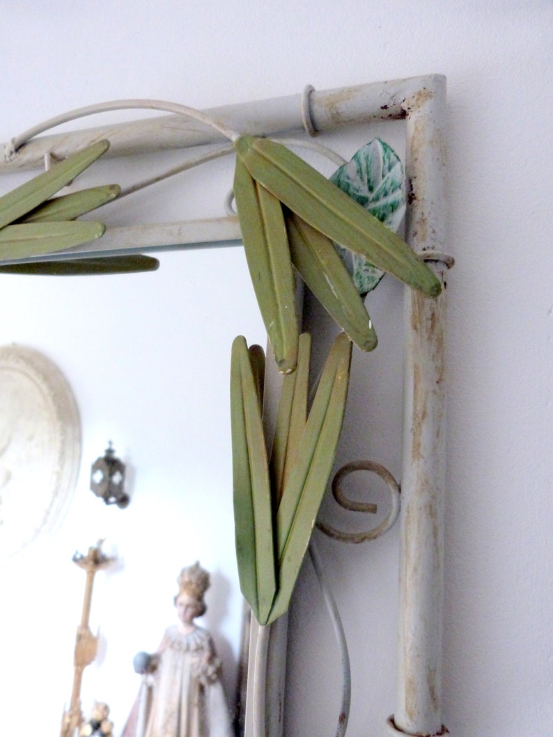 May include: A white metal mirror with a decorative design of green leaves. The mirror has a distressed white finish and is framed with a metal frame.
