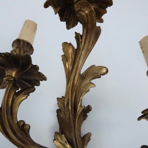 K&ouml;nnte beinhalten: Eine goldfarbene Wandleuchte mit drei Gl&uuml;hbirnen. Die Leuchte ist aus verziertem Metall mit einem floralen Design.