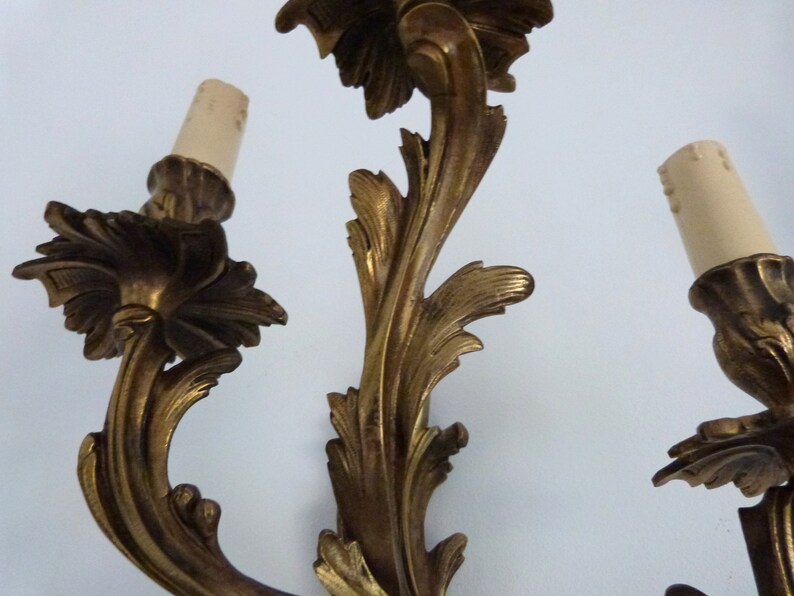 K&ouml;nnte beinhalten: Eine goldfarbene Wandleuchte mit drei Gl&uuml;hbirnen. Die Leuchte ist aus verziertem Metall mit einem floralen Design.