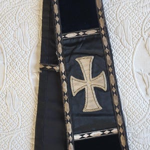May include: A black liturgical stole with a large, embroidered cross in the center. The stole has a velvet panel and is trimmed with gold and silver decorative elements. The bottom edge features a fringe.