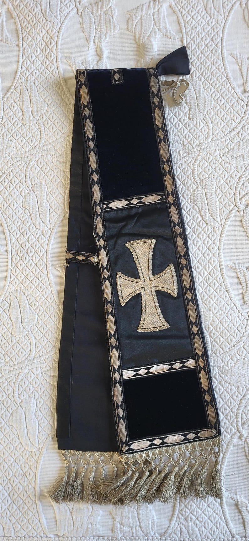 May include: A black liturgical stole with a large, embroidered cross in the center. The stole has a velvet panel and is trimmed with gold and silver decorative elements. The bottom edge features a fringe.