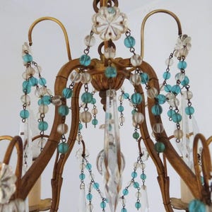 May include: Close-up of a vintage chandelier with a gold-colored frame. The chandelier is adorned with clear crystal pendants, turquoise and clear glass beads, and decorative floral accents. The design is ornate and elegant.