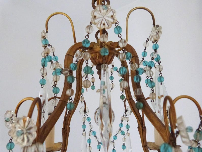 May include: Close-up of a vintage chandelier with a gold-colored frame. The chandelier is adorned with clear crystal pendants, turquoise and clear glass beads, and decorative floral accents. The design is ornate and elegant.