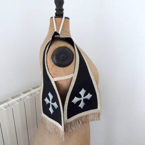 Może przedstawiać: Czarna i biała stuła religijna z motywem krzyża. Stuła ma kremowe frędzle i wykończenia. Przedmiot jest prezentowany na zabytkowym manekinie.