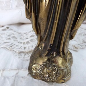 Op de afbeelding: Een gouden metalen beeld van een vrouw in een lange jurk. Het beeld staat op een voet met een decoratief ontwerp.