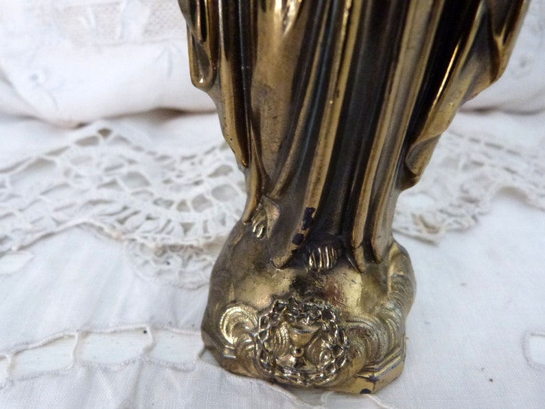 Op de afbeelding: Een gouden metalen beeld van een vrouw in een lange jurk. Het beeld staat op een voet met een decoratief ontwerp.