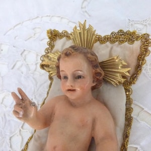 May include: A vintage figurine of a baby with light skin, brown hair, and a serene expression. The baby is posed with one hand raised in a peace sign. The figurine rests on a cream-colored pillow with gold trim and a gold halo.