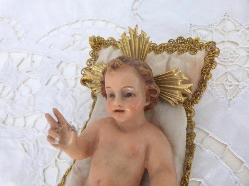 May include: A vintage figurine of a baby with light skin, brown hair, and a serene expression. The baby is posed with one hand raised in a peace sign. The figurine rests on a cream-colored pillow with gold trim and a gold halo.