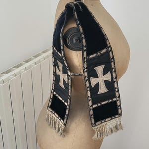 May include: A black and cream-colored religious stole with a cross design. The stole is made of a dark fabric with cream-colored crosses and trim. The bottom of the stole has a cream-colored fringe.
