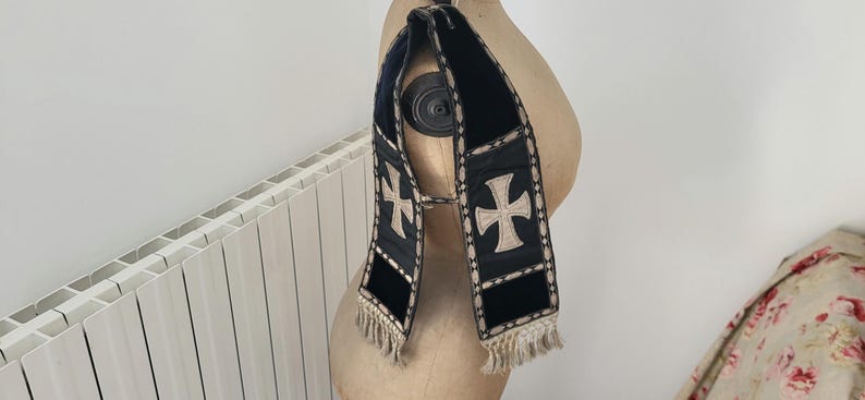 May include: A black and cream-colored religious stole with a cross design. The stole is made of a dark fabric with cream-colored crosses and trim. The bottom of the stole has a cream-colored fringe.