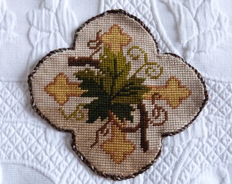 May include: A brown, cream, and green embroidered cross-shaped needlepoint with a floral design. The design features leaves and flowers in shades of green, brown, and yellow.