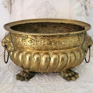 Puede incluir: Una jardinera de latón vintage con acabado dorado. La jardinera presenta asas con cabeza de león y patas de garra. El borde y el cuerpo están adornados con patrones en relieve. Una pieza decorativa.