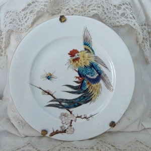 May include: A white porcelain plate with a gold rim features a hand-painted design of a colorful bird with blue, yellow, and red feathers perched on a branch with pink blossoms. A small bee is also painted on the plate.