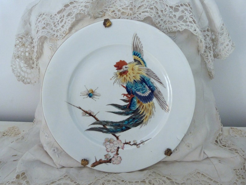 May include: A white porcelain plate with a gold rim features a hand-painted design of a colorful bird with blue, yellow, and red feathers perched on a branch with pink blossoms. A small bee is also painted on the plate.