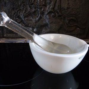 May include: A white ceramic mortar and pestle set. The mortar is round and has a slightly flared lip. The pestle is made of clear glass and has a rounded end.
