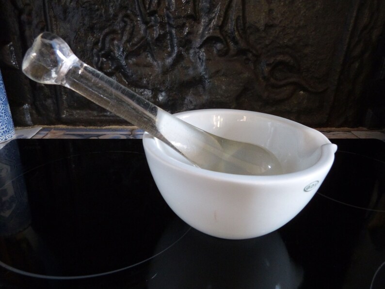 May include: A white ceramic mortar and pestle set. The mortar is round and has a slightly flared lip. The pestle is made of clear glass and has a rounded end.