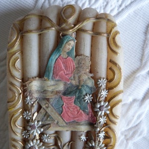 May include: A white candle with a gold trim and a picture of Mary and baby Jesus in a manger. The candle is decorated with silver flowers.