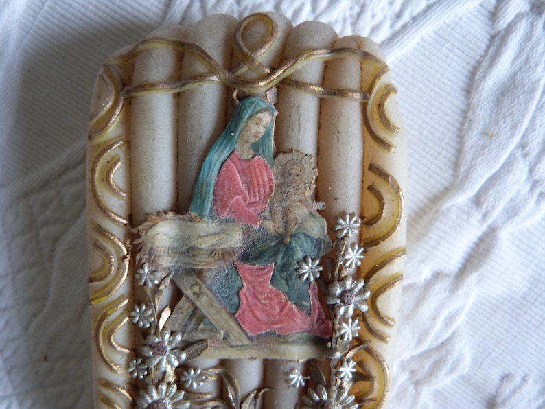 May include: A white candle with a gold trim and a picture of Mary and baby Jesus in a manger. The candle is decorated with silver flowers.