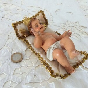 May include: A vintage figurine of a baby, lying on a white pillow with gold trim. The baby has light skin and is wearing a white diaper. A gold halo is behind the baby's head. A coin is next to the pillow.