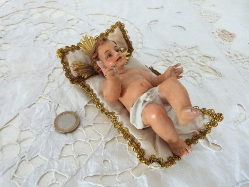 May include: A vintage figurine of a baby, lying on a white pillow with gold trim. The baby has light skin and is wearing a white diaper. A gold halo is behind the baby's head. A coin is next to the pillow.