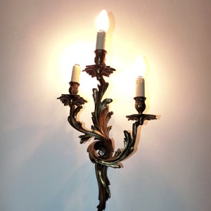 K&ouml;nnte beinhalten: Eine dreiarmige Wandleuchte mit goldener Oberfl&auml;che und verzierten Schn&ouml;rkeln. Jeder Arm hat eine Kerzen-f&ouml;rmige Gl&uuml;hbirne.