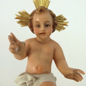May include: A detailed figurine of a baby with a golden halo. The baby has a light skin tone, rosy cheeks, and is wearing a white cloth. The halo is made of gold-colored rays.