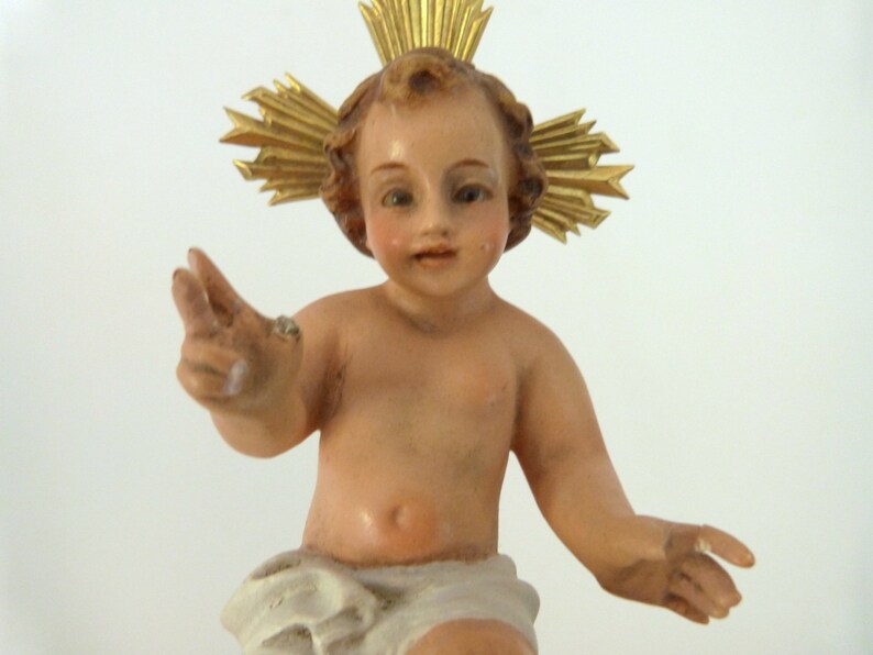 May include: A detailed figurine of a baby with a golden halo. The baby has a light skin tone, rosy cheeks, and is wearing a white cloth. The halo is made of gold-colored rays.
