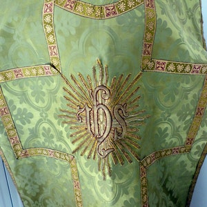 May include: A green liturgical vestment with a floral pattern and gold trim. The center features an embroidered IHS monogram surrounded by sun rays. The vestment has a detailed design with a decorative border.