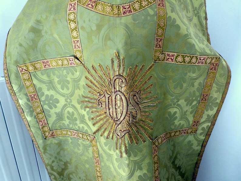 May include: A green liturgical vestment with a floral pattern and gold trim. The center features an embroidered IHS monogram surrounded by sun rays. The vestment has a detailed design with a decorative border.