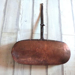 May include: Antique copper bed warmer with a long, dark metal handle. The copper warming pan has an oval shape with decorative circular patterns. The handle has a hinged mechanism for folding.