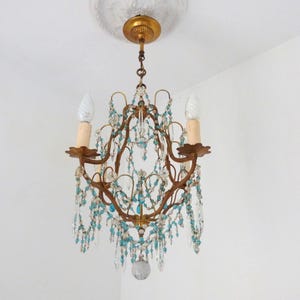 May include: An antique chandelier with a bronze frame, adorned with clear and turquoise crystal beads. The chandelier has two light fixtures with off-white candle-shaped bulbs. The fixture is suspended from a decorative ceiling mount.