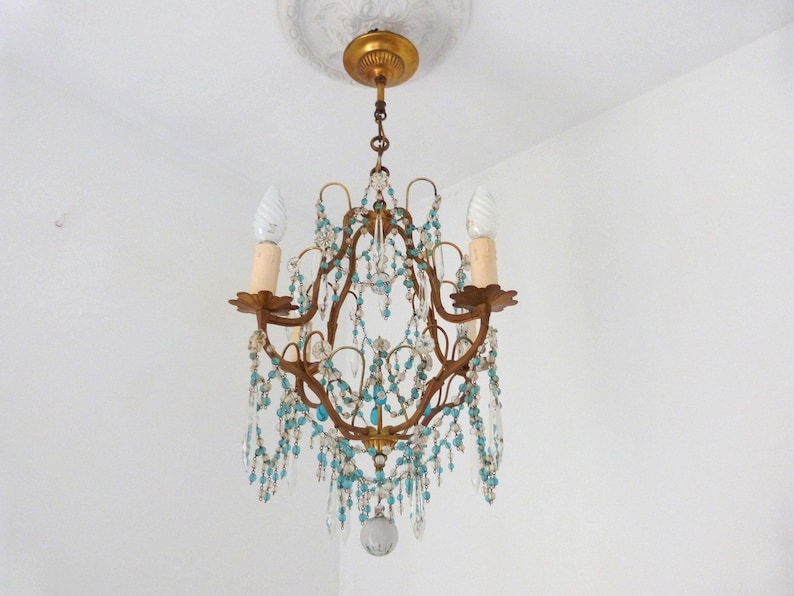 May include: An antique chandelier with a bronze frame, adorned with clear and turquoise crystal beads. The chandelier has two light fixtures with off-white candle-shaped bulbs. The fixture is suspended from a decorative ceiling mount.