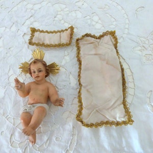 May include: A detailed figurine of a baby with a golden halo, wearing a diaper. Two fabric pieces with gold trim are also present. The items are arranged on a white, lace-like surface. The baby figurine is the focal point.