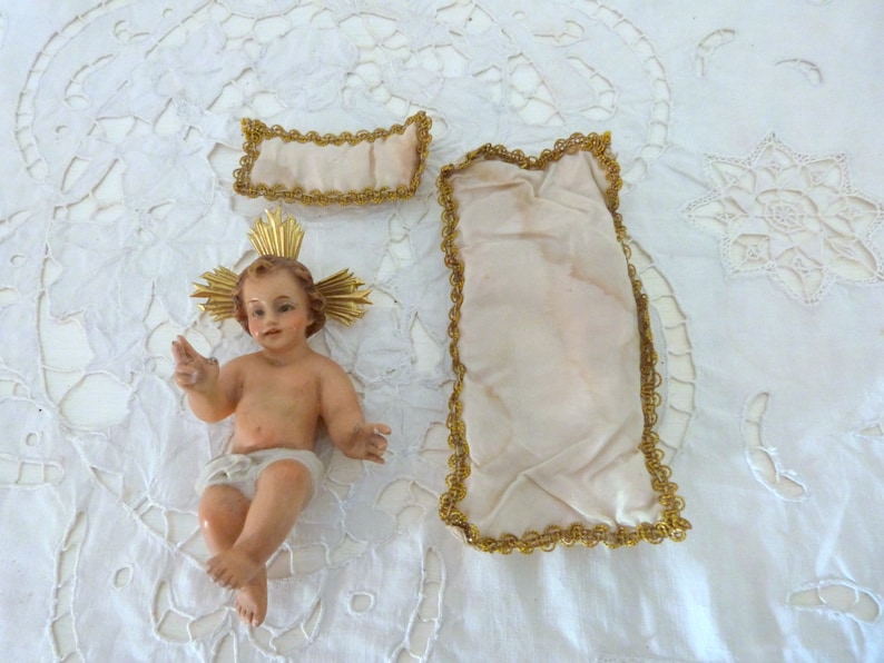 May include: A detailed figurine of a baby with a golden halo, wearing a diaper. Two fabric pieces with gold trim are also present. The items are arranged on a white, lace-like surface. The baby figurine is the focal point.