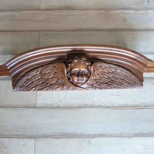 Puede incluir: Elemento arquitectónico de madera tallada que presenta la cara de un ángel con alas detalladas. La pieza es arqueada y tiene un acabado marrón cálido. La cara del ángel está centrada, con las alas extendiéndose hacia afuera.