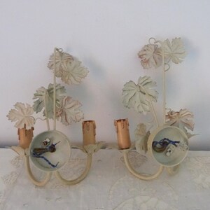 May include: A pair of white metal wall sconces with a grapevine design. Each sconce has two candle holders and a circular base.