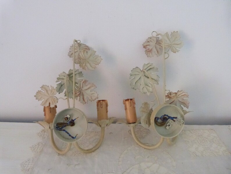 May include: A pair of white metal wall sconces with a grapevine design. Each sconce has two candle holders and a circular base.