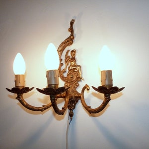 K&ouml;nnte beinhalten: Eine goldfarbene Wandleuchte mit drei Gl&uuml;hbirnen. Die Leuchte hat ein dekoratives Design mit einer Figur einer Frau, die auf einem Thron sitzt.