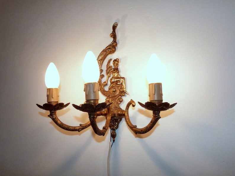 K&ouml;nnte beinhalten: Eine goldfarbene Wandleuchte mit drei Gl&uuml;hbirnen. Die Leuchte hat ein dekoratives Design mit einer Figur einer Frau, die auf einem Thron sitzt.