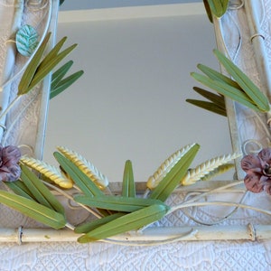 May include: A white metal mirror with a floral design. The mirror is decorated with green leaves, yellow wheat stalks, and pink flowers.