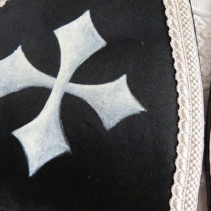 Może przedstawiać: Czarny element odzieży z dużym białym krzyżem. Krzyż jest pomalowany z efektem tekstury. Krawędź odzieży jest obszyta białą koronką. Og&oacute;lny styl sugeruje motyw religijny lub gotycki.