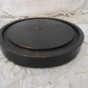 Peut inclure: Un objet rond en bois brun foncé, avec un dessus plat et une base légèrement surélevée. Il est posé sur une surface blanche recouverte de dentelle, évoquant un style vintage. Le dessus présente un petit trou central.