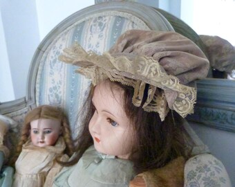 May include: Antique porcelain doll with brown hair and blue eyes, wearing a light green dress and a lace-trimmed bonnet. The doll is seated in a vintage chair with a floral pattern.