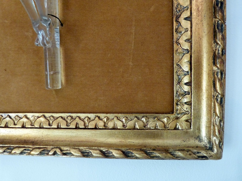 May include: A gold-colored picture frame with a brown velvet mat and a glass object mounted inside. The frame has a decorative, ornate design.