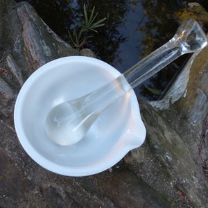 May include: A white ceramic mortar and pestle with a clear glass pestle. The mortar and pestle are sitting on a wooden surface.