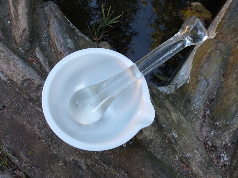 May include: A white ceramic mortar and pestle with a clear glass pestle. The mortar and pestle are sitting on a wooden surface.