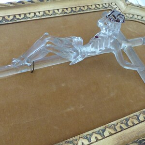 May include: A glass crucifix with a clear, translucent finish. The figure is lying on a cross with a red stain on the chest. The crucifix is displayed in a gold-colored frame with ornate detailing.