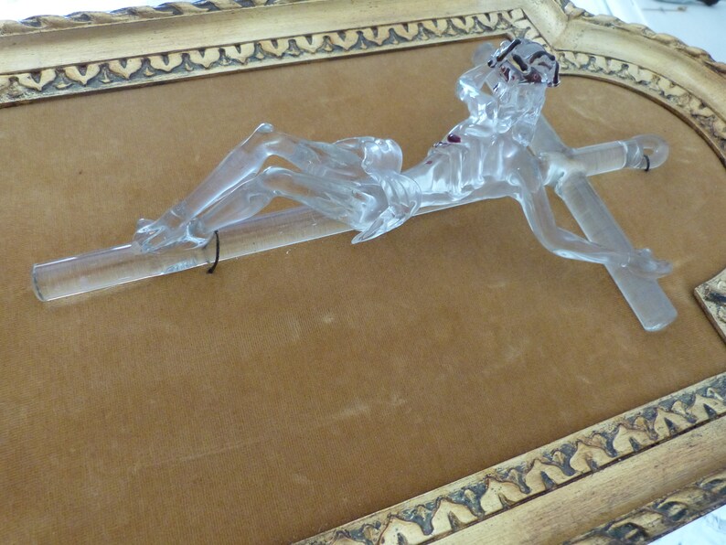 May include: A glass crucifix with a clear, translucent finish. The figure is lying on a cross with a red stain on the chest. The crucifix is displayed in a gold-colored frame with ornate detailing.